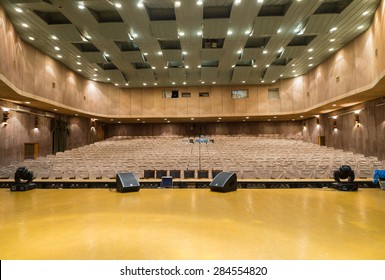 View Of Empty Seats With Seat Covers Looking Toward Back Of Theater From Empty Stage In Well Lit Venue