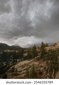 View from Estes Park towards the dramatic peaks of Rocky Mountain National Park under ominous storm clouds. A serene mountain scene with lush evergreen trees, rocky terrain, and distant fog-covered su