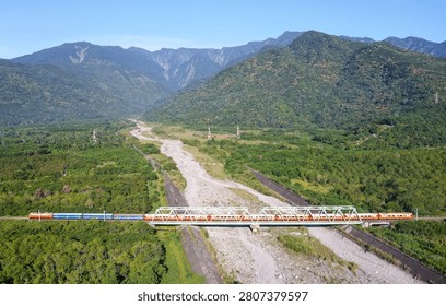 View of an express train dashing through an iron bridge over the dried riverbed of Kanon River on a sunny day with beautiful mountains under blue clear sky in background in Guangfu Township, Hualien County, Taiwan.
This magnificent wide-angle aerial photograph captures the breathtaking natural splendor and rhythmic infrastructure of eastern Taiwan's Hualien County (花蓮縣). The composition centers on a long passenger train operated by the Taiwan Railway Corporation (台鐵) as it glides horizontally across the frame. The train is traversing the Jianongnong River Bridge (加濃濃溪橋), a distinct silver-white steel truss railway bridge that spans across the expansive, gravel-strewn dry riverbed of the Jianongnong River (a tributary of the Guangfu ). The landscape layout is a striking contrast of geometric order and wild nature. Flanking the grey riverbed are endless miles of lush, vibrant emerald-green forests and agricultural fields characteristic of the Central Mountain Valley. In the background, the horizon is dominated by the majestic, towering ridge lines of the Central Mountain Range (中央山脈), draped in deep forest greens and climbing toward a flawless, clear blue sky. The morning sun illuminates the side of the mountain and the train cars, creating an incredibly crisp, bright, and harmonious depiction of rural Taiwan.
The location visible in this frame represents a crucial segment of the Taitung Line (台東線), which serves as the transport backbone of eastern Taiwan. The Jianongnong viaduct sits on the border of Guangfu Township (光復鄉), an area steeped in the history of the indigenous Amis (阿美族) people. The river's unique name, Jianongnong (加濃濃), is derived from the original Amis place name Kanana, which refers to a traditional type of woven bamboo basket once used by the tribe for fishing and collecting wild crops in this exact valley. The Taitung Line originally opened during the early 20th century as a narrow-gauge railway before being progressively widened and rerouted.
