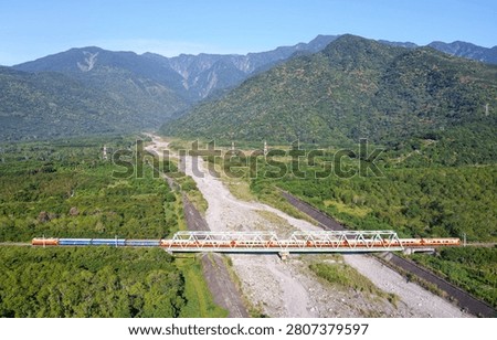 View of an express train dashing through an iron bridge over the dried riverbed of Kanon River on a sunny day with beautiful mountains under blue clear sky in background in Guangfu Township, Hualien County, Taiwan.
This magnificent wide-angle aerial photograph captures the breathtaking natural splendor and rhythmic infrastructure of eastern Taiwan's Hualien County (花蓮縣). The composition centers on a long passenger train operated by the Taiwan Railway Corporation (台鐵) as it glides horizontally across the frame. The train is traversing the Jianongnong River Bridge (加濃濃溪橋), a distinct silver-white steel truss railway bridge that spans across the expansive, gravel-strewn dry riverbed of the Jianongnong River (a tributary of the Guangfu ). The landscape layout is a striking contrast of geometric order and wild nature. Flanking the grey riverbed are endless miles of lush, vibrant emerald-green forests and agricultural fields characteristic of the Central Mountain Valley. In the background, the horizon is dominated by the majestic, towering ridge lines of the Central Mountain Range (中央山脈), draped in deep forest greens and climbing toward a flawless, clear blue sky. The morning sun illuminates the side of the mountain and the train cars, creating an incredibly crisp, bright, and harmonious depiction of rural Taiwan.
The location visible in this frame represents a crucial segment of the Taitung Line (台東線), which serves as the transport backbone of eastern Taiwan. The Jianongnong viaduct sits on the border of Guangfu Township (光復鄉), an area steeped in the history of the indigenous Amis (阿美族) people. The river's unique name, Jianongnong (加濃濃), is derived from the original Amis place name Kanana, which refers to a traditional type of woven bamboo basket once used by the tribe for fishing and collecting wild crops in this exact valley. The Taitung Line originally opened during the early 20th century as a narrow-gauge railway before being progressively widened and rerouted.