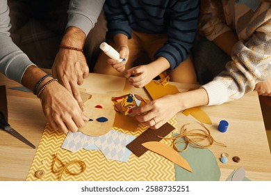 Vista de los miembros de la familia creando manualidades de papel hechas a mano juntos, centrándose en las manos que trabajan con pegamento y materiales de papel de colores colocados en la mesa de madera