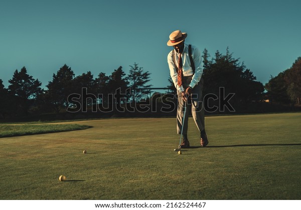 View of a fancy mature African guy in an elegant\
retro outfit with a straw hat and a tie hitting the ball with a\
club while standing on the lawn of the private golf club on a warm\
sunny evening