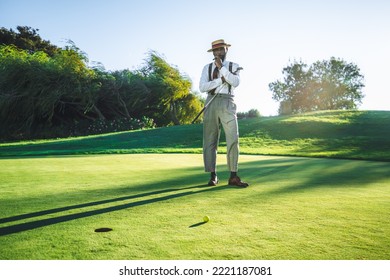 View of a fashionable handsome bearded black senior with a sigar, in straw hat and tailored outfit standing on a green golf field with a club in his hand; a yellow golf ball and a hole in front of him - Powered by Shutterstock