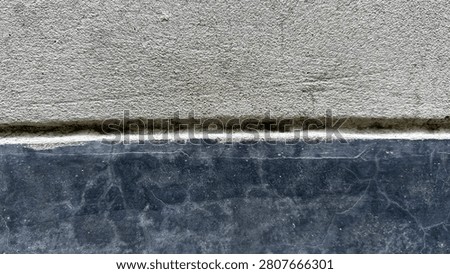 A view featuring two distinct horizontal sections of a wall. The upper portion consists of a rough, light gray concrete surface with a porous, grainy texture. The lower portion is a dark, smooth, polished stone slab with subtle, faint white veining or hairline cracks. A clear, horizontal seam or mortar line separates the two contrasting creating a sharp geometric division across the frame. The lighting is even, highlighting the difference in texture between the two surfaces.