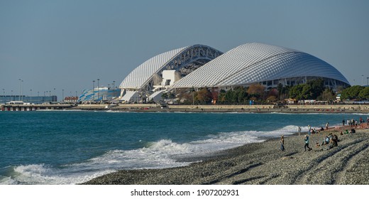 View Of Fisht Stadium From Olympic Embankment In Sirius. Blue Waves And Gray Pebbles Of Black Sea Coast. People Resting On Sunny Winter Day.  Sochi, Russia - December 08, 2020