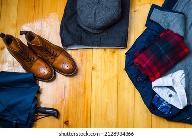 View Flat Lay of Tan Brogues Boots With Umbrella, Jumper,Checked Winter Scarf, Cap, Herringbone Trousers, White Shirt and Raincoat on Wooden Surface Background. Horizontal Image