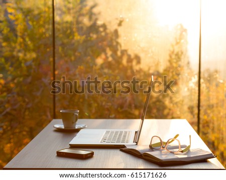 View of Freelancer working Place at wooden Table with Computer Coffee Mug Notepad Telephone and Glasses at Morning in Forest Villa