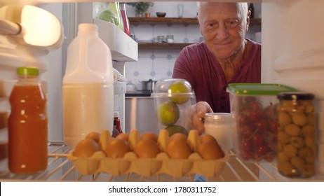 View From Fridge Of Senior Man Unpacking Paper Bags With Groceries And Putting Food On Shelves In Refrigerator In Kitchen At Home. Husband Doing House Chores