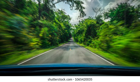 View From Front Of Blue Car On Asphalt Road And Speed Motion Blur On Highway In Summer With Green Trees Forest At Countryside. Road Trip Travel On Green Season. Car With Blur Motion And Blue Sky.