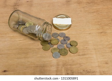 View frop top of coins spilling from a glass jar attached with empty white paper on the lid over wooden background. Free copy space. Financial concept.