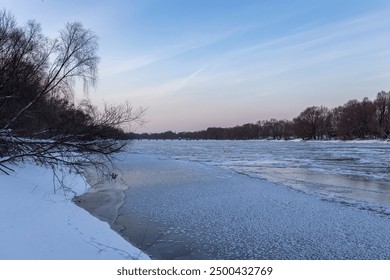 A view of a frozen river, with snow-covered banks, amidst a peaceful, twilight sky. Bare trees line the river's edge, adding to the wintery scene. - Powered by Shutterstock