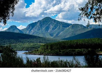 View of Garden Route Dam with forest trees and dam appearing the foreground and Outeniqua Mountain Range appearing in the background, clouds overpowering the blue sky. - Shutterstock ID 2166382427
