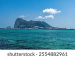 View of Gibraltar Rock from La Linea de la Concepcion coastline. In the foreground, turquoise harbor water with city and mountains in the background.