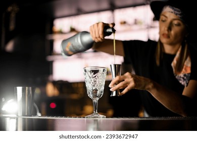 View of girl bartender at bar counter holding bottle with syrup and pours it to steel jigger for making cocktail. Woman bartender creates tasty alcoholic drink - Powered by Shutterstock