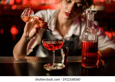 view of glass with bright red alcoholic drink to which woman bartender carefully adds ingredient from small bottle