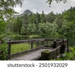 View of Glencoe Lochan, Scotland, surrounded by forest with cloudy sky, wooden jetty in foreground and and mountain, Pap of Glencoe, in background