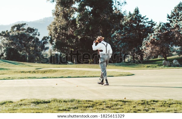 View of a golf field on a sunny windy day with an\
elegant retrosexual black guy in a hat, white shirt, and suspender\
trousers, he is walking to a ball on the grass to hit it towards\
the closest hole