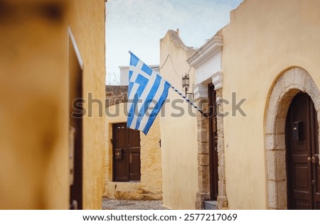 View of Greek flag waving in air serene Mediterranean vibe. beautiful Greek islands series, Narrow cobblestone street lined with charm in downtown Rhodes , Greece. Idyllic stone alley