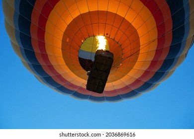view from the ground of colorful hot air balloon with large wicker passenger basket rising up in the blue sky using burner fire flame