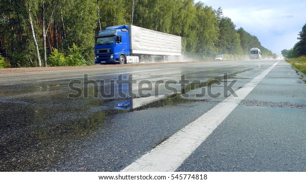View from ground of\
traffic on wet road, highway with mist splash after rain with green\
trees on a roadside on background. Trucks are passing by camera in\
perspective