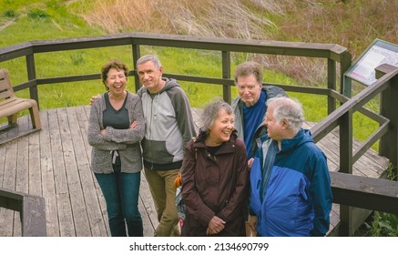 View Of Group Of Older Friends Standing On Wooden Deck Looking At Each Other And Laughing On A Walk In Midwestern Park 