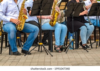 View Of A Group Of People From A Musical Brass Band Who Are Sitting On Chairs And Playing Music, Jazz On Golden Copper Pipes, Sitting Outside In The Open Air. Photos Without Permission Of Models