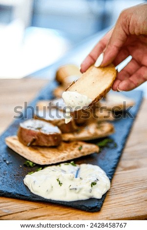 View hand dipping potato chips garnished with garlic, rosemary and thyme into yogurt garlic chive dip sauce on black plate and wooden table. Meal with meat, potatos and bread.