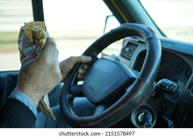 View Of A Hand Of A Man Holding Food And Driving At The Same Time. Dangerous Driving.