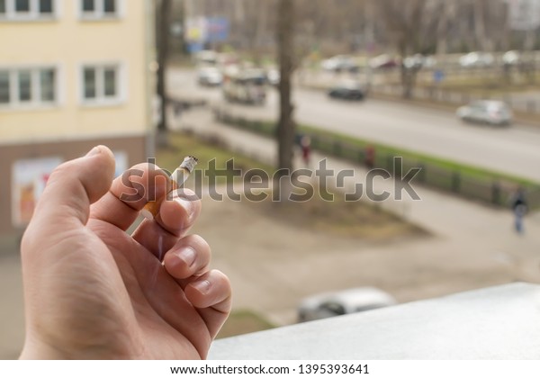 View of the hand of a Smoking man with a\
Smoking cigarette from the window of the apartment into the yard\
and the street with passing cars on the\
road