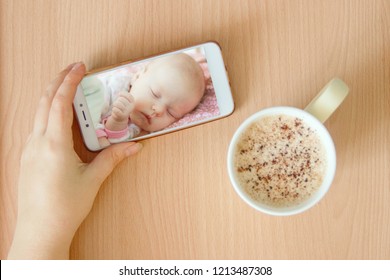 View Handheld Color Video Baby Monitor. Female Hands Are Holding A Smartphone With A Baby Monitor App. Near Hot Drink.