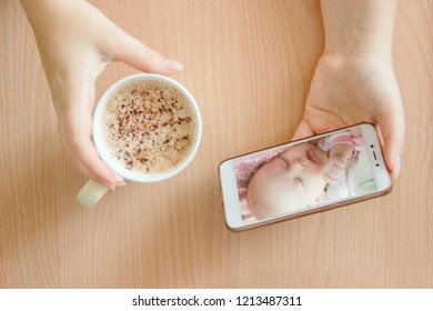 View Handheld Color Video Baby Monitor. Female Hands Are Holding A Smartphone With A Baby Monitor App. Near Hot Drink.