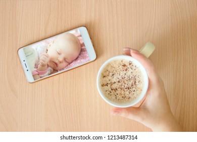 View Handheld Color Video Baby Monitor. Female Hands Are Holding A Smartphone With A Baby Monitor App. Near Hot Drink.