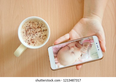 View Handheld Color Video Baby Monitor. Female Hands Are Holding A Smartphone With A Baby Monitor App. Near Hot Drink.