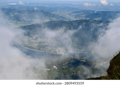 A view from high above, looking down through thick white clouds onto a long, winding body of water, a small town, and a green valley floor.