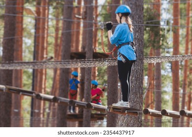 View of high ropes course, kids of climbing in amusement acitivity rope park, passing obstacles and zip line on heights, children teenagers in equipment gear between the trees on heights, summer day - Powered by Shutterstock