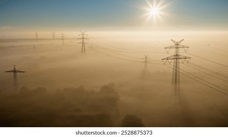 View of high-voltage power lines stretching through a mist-covered landscape at sunrise, with golden light casting long shadows and illuminating the fog-draped fields. - Powered by Shutterstock