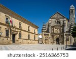 View of the historic Capilla del Salvador alongside the Renaissance Parador de Turismo in Ubeda, Jaen, Andalucia. The landmarks are set against a clear blue sky.