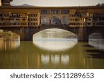 View of the historic Ponte Vecchio bridge reflecting on the Arno River in Florence, Italy, during sunset.