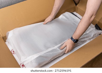 View of human hands carefully taking a new electronic item in its white protective packaging from a brown cardboard delivery box, showcasing online commerce and anticipation