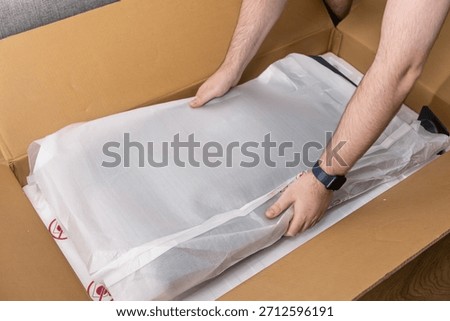 View of human hands carefully taking a new electronic item in its white protective packaging from a brown cardboard delivery box, showcasing online commerce and anticipation