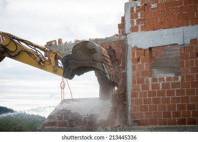 View Of The Hydraulic Arm And Bucket Of A Large Heavy Duty Backhoe Demolishing A Brick House Breaking Down The Exterior Wall For Removal.