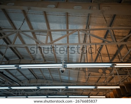 A view of an industrial ceiling with a security camera and a row of fluorescent lights mounted below the steel truss structure and reflective insulation, with warm light on the right.
