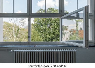 A view from inside a bright room looking out through a large, white-framed window with one pane swung open to let in fresh air. Outside, lush green trees and a blue sky with clouds are visible, while 