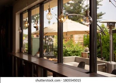 A view from inside a cafe setting, looking through a wall of windows towards a comfortable isolated patio area, featuring vintage hanging light bulbs. - Powered by Shutterstock