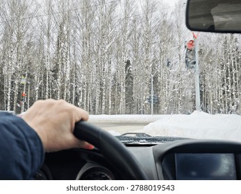 View from inside a car at an intersection with a traffic light during winter with snowy trees in the background and a hand on the wheel.