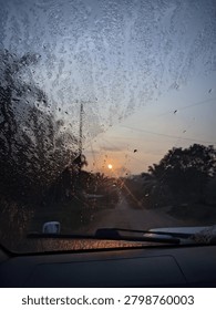 View from inside a car looking through a dusty or wet windshield at a sunset over a rural road. Silhouettes of trees and  owner lines frame the glowing sun on the horizon. A moody travel concept capturing the atmosphere of a road trip at dusk.
Vertical composition with textured fore ground. 