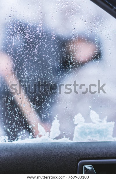 View from inside of car on man dressed in jacket\
cleaning snow around his\
car.