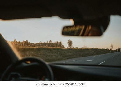 View from inside a car onto a road running through a forest. The sun is setting, creating a warm glow. The steering wheel, side mirror and a bit of asphalt road are visible. High quality photo - Powered by Shutterstock