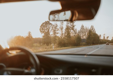 View from inside a car onto a road running through a forest. The sun is setting, creating a warm glow. The steering wheel, side mirror and a bit of asphalt road are visible. High quality photo - Powered by Shutterstock