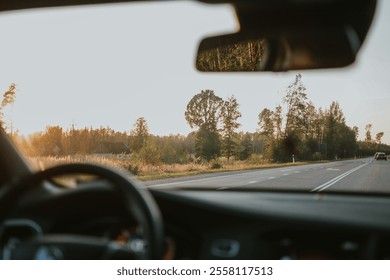 View from inside a car onto a road running through a forest. The sun is setting, creating a warm glow. The steering wheel, side mirror and a bit of asphalt road are visible. High quality photo - Powered by Shutterstock